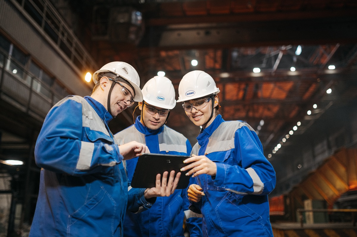 three factory workers wearing white hard hats, using a tablet